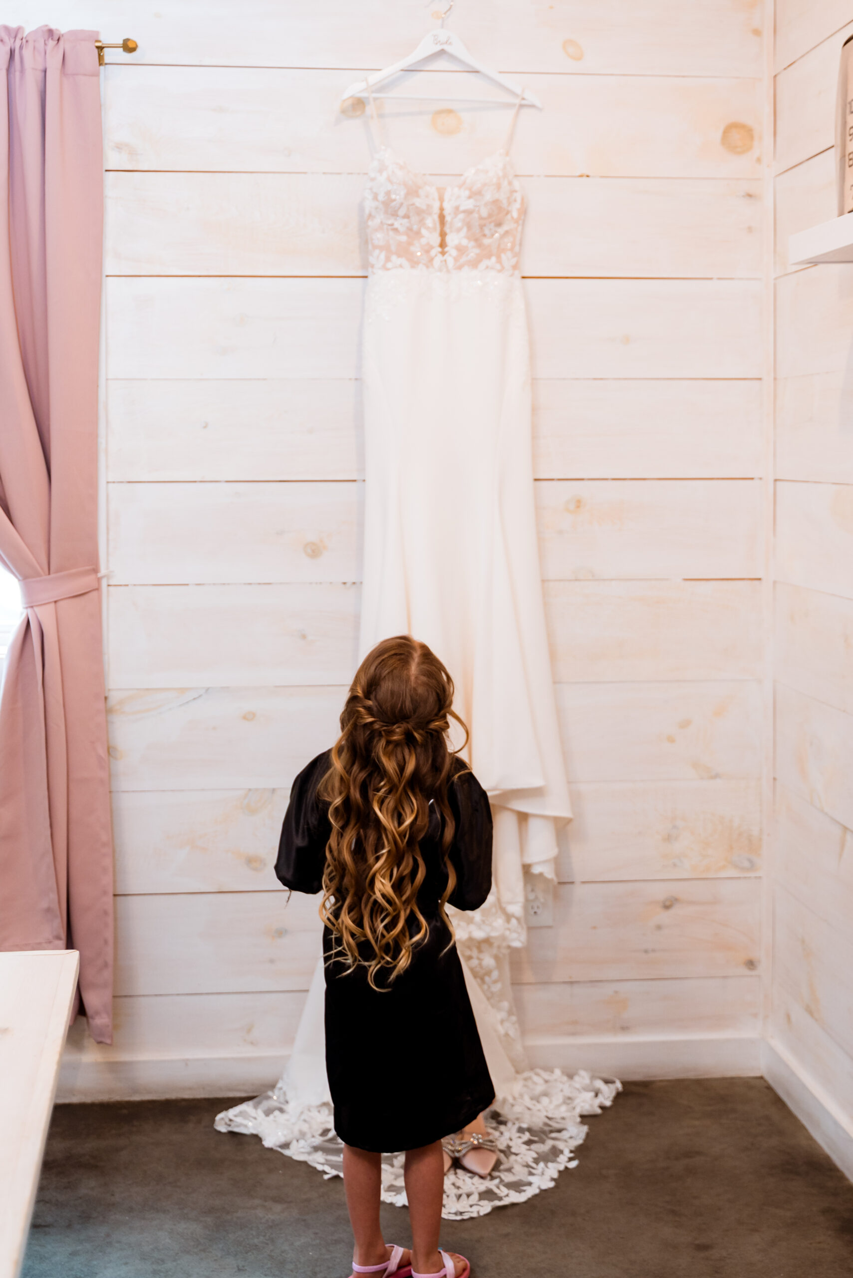 Lace wedding dress hanging on a white shiplap wall as a flower girl looks up at it in the bridal suite at Stonehurst at Hampton Valley in Hampton CT