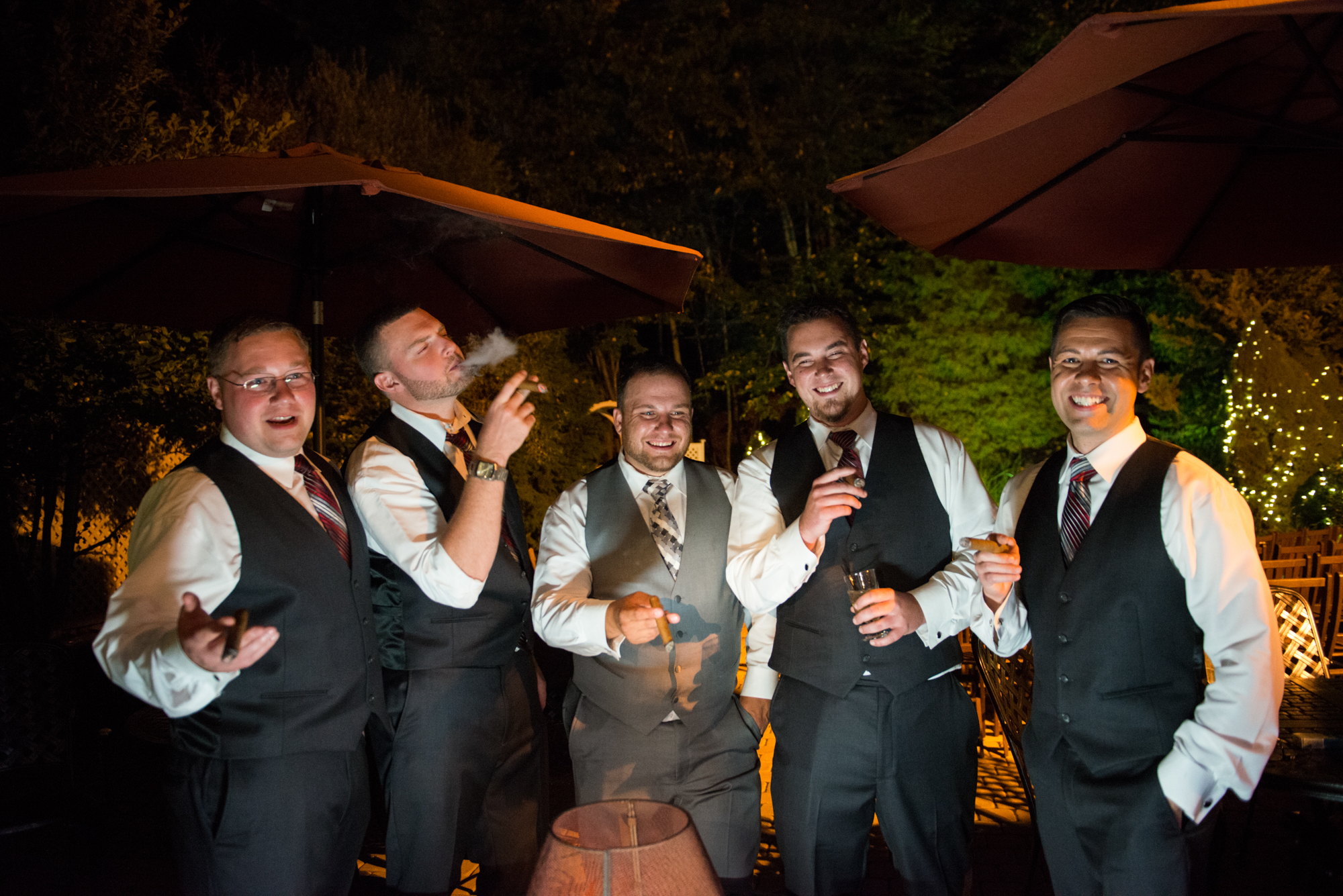 Groomsmen laughing and enjoying cigars on the outdoor patio at night during a wedding reception at The Woodwinds in Branford CT