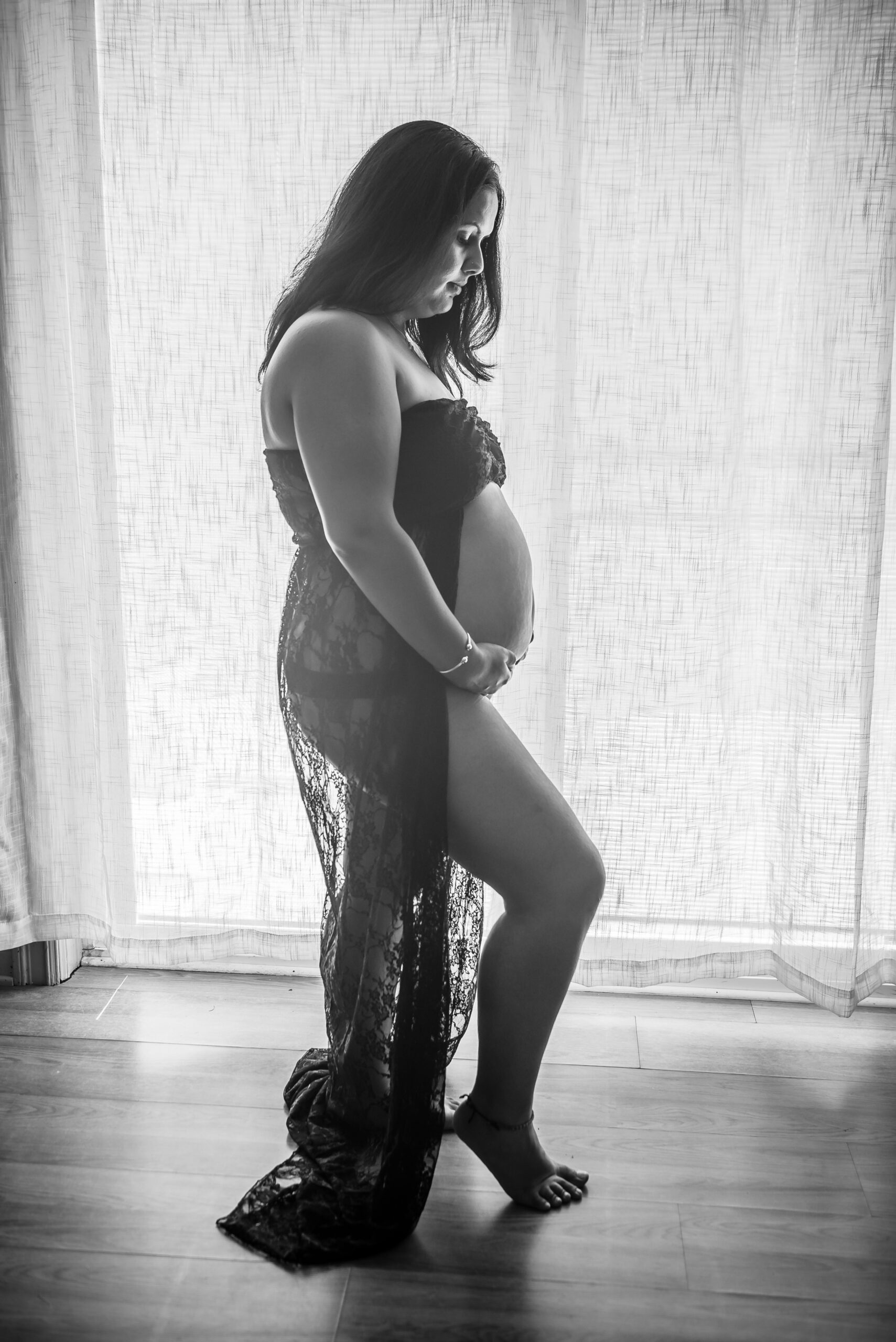 Black and white maternity portrait of a woman in a sheer lace gown cradling her belly beside a sunlit window in Connecticut