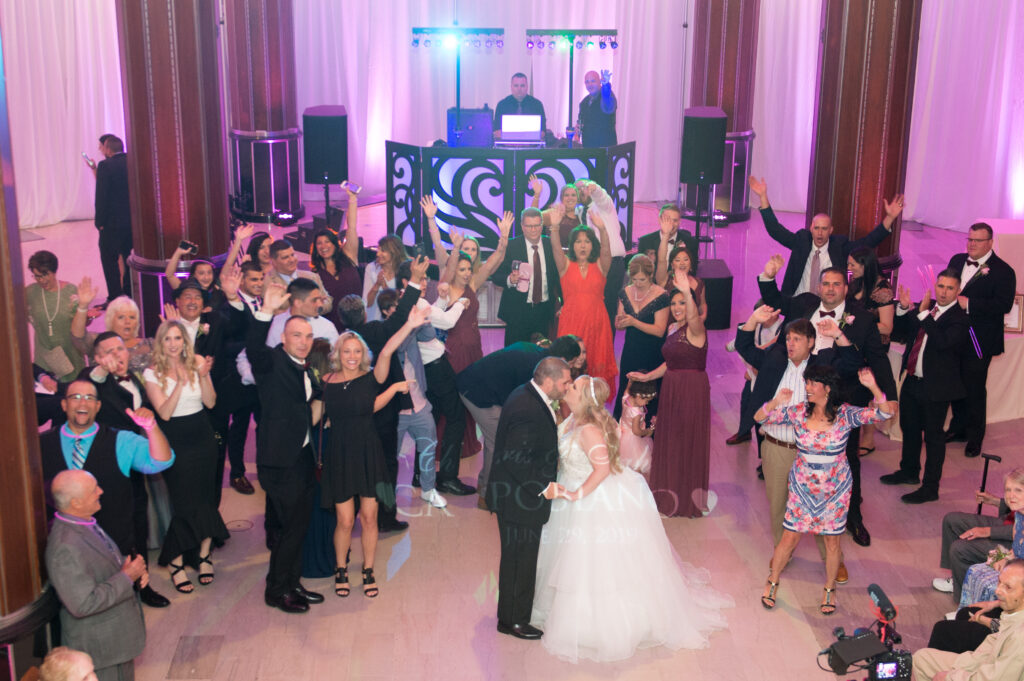Bride and groom kissing surrounded by cheering guests with two DJs at the ornate heart booth and pink and purple uplighting at the G. Fox Ballroom in Hartford CT