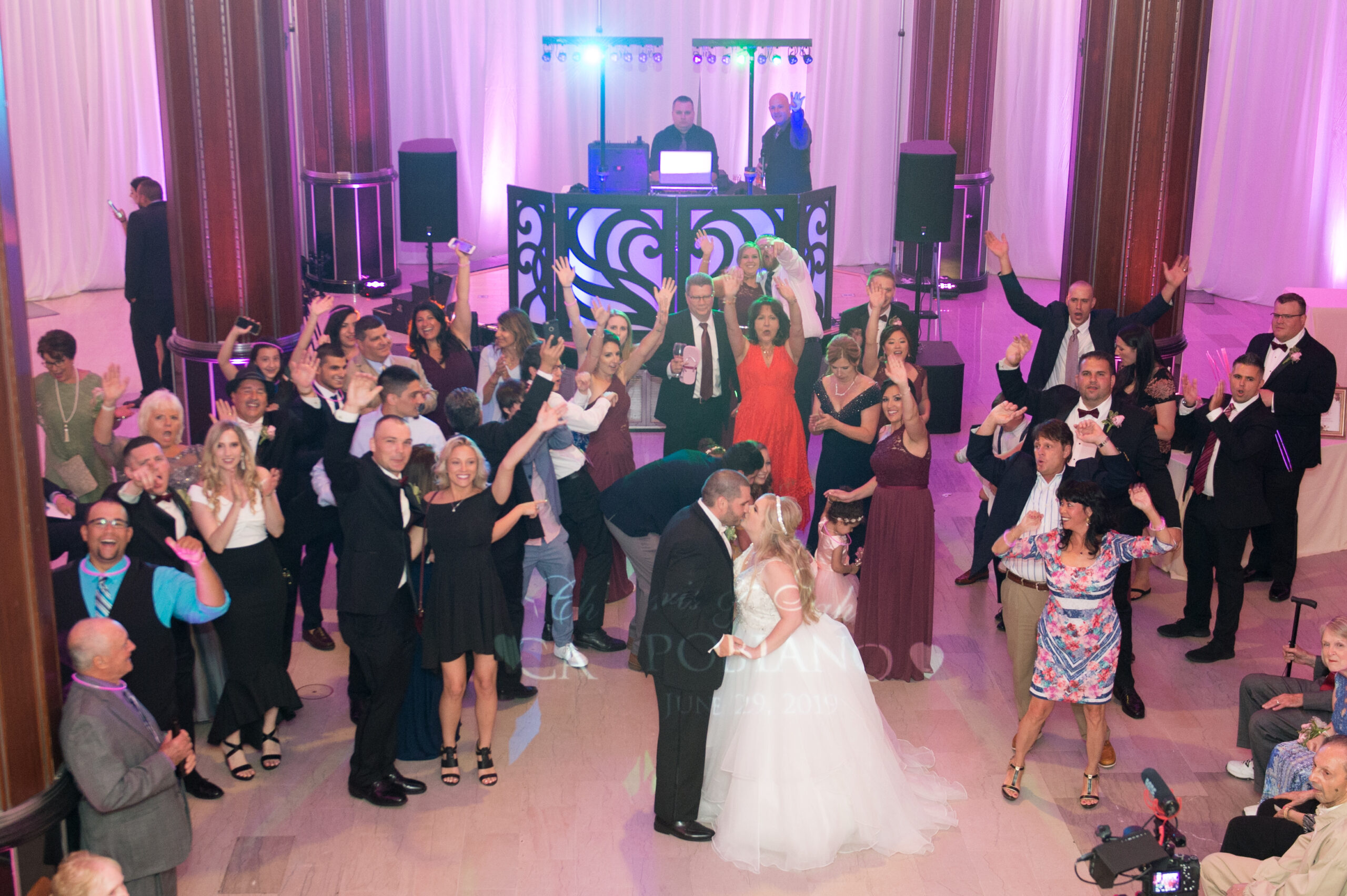 Bride and groom kissing surrounded by cheering guests with two DJs at the ornate heart booth and pink and purple uplighting at the G. Fox Ballroom in Hartford CT
