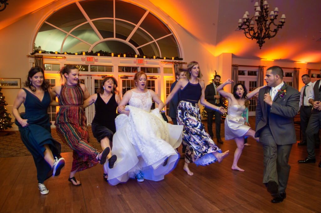 Bride holding her gown and kicking up her sneakers in a line dance with guests under warm uplighting at Riverhouse at Goodspeed Station in East Haddam CT