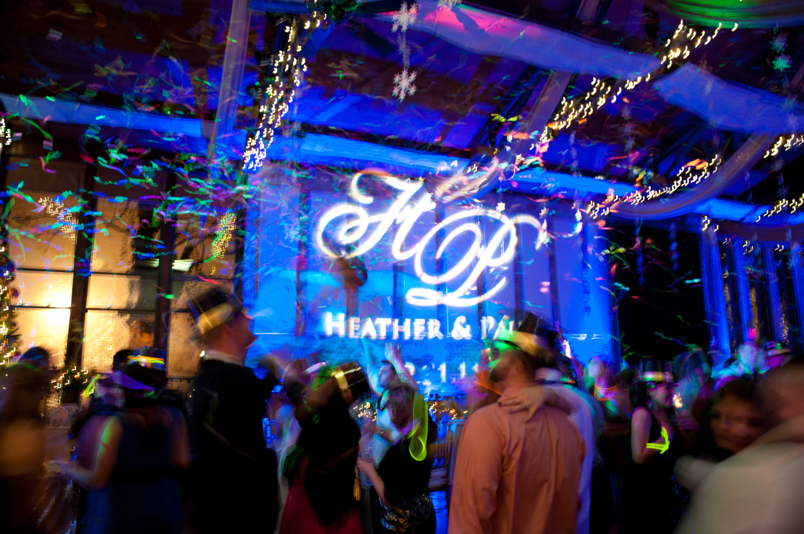 Custom monogram projection glowing on a blue-lit wall as guests celebrate with confetti and party hats at a Pond House Cafe wedding in West Hartford CT