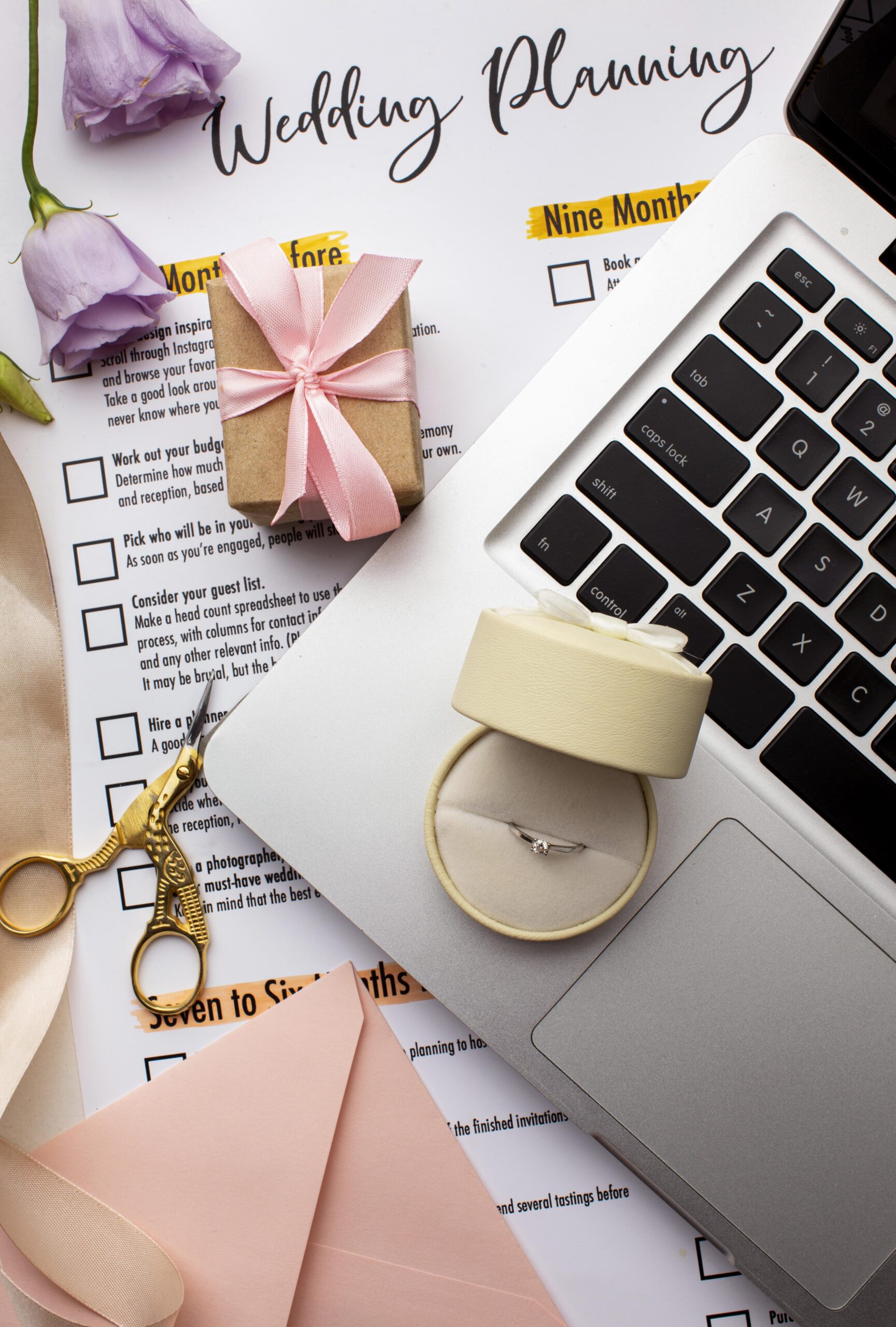 Wedding planning checklist with engagement ring in box, pink envelopes, laptop, and gold scissors styled on a flat-lay