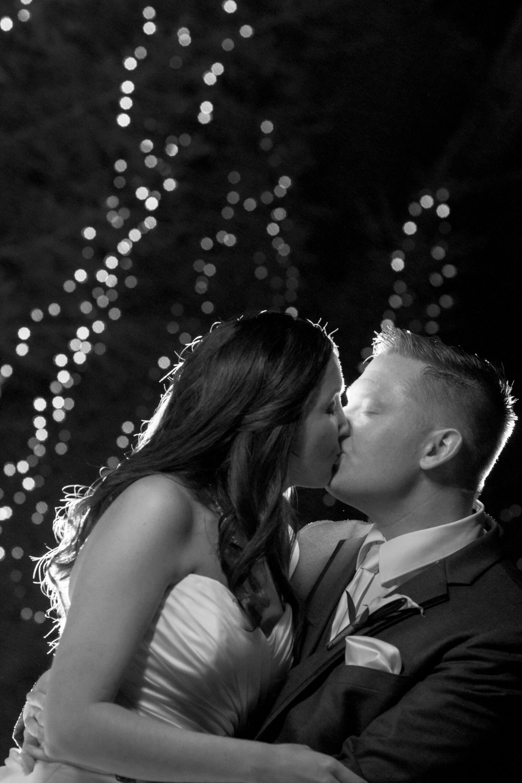 Bride and groom kissing at night with twinkling string lights glowing behind them at The Woodwinds in Branford CT, black and white